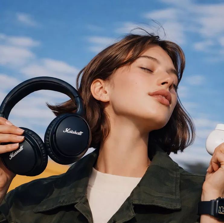A woman holds headphones in one hand and a cup of coffee in the other, smiling as she enjoys her morning routine